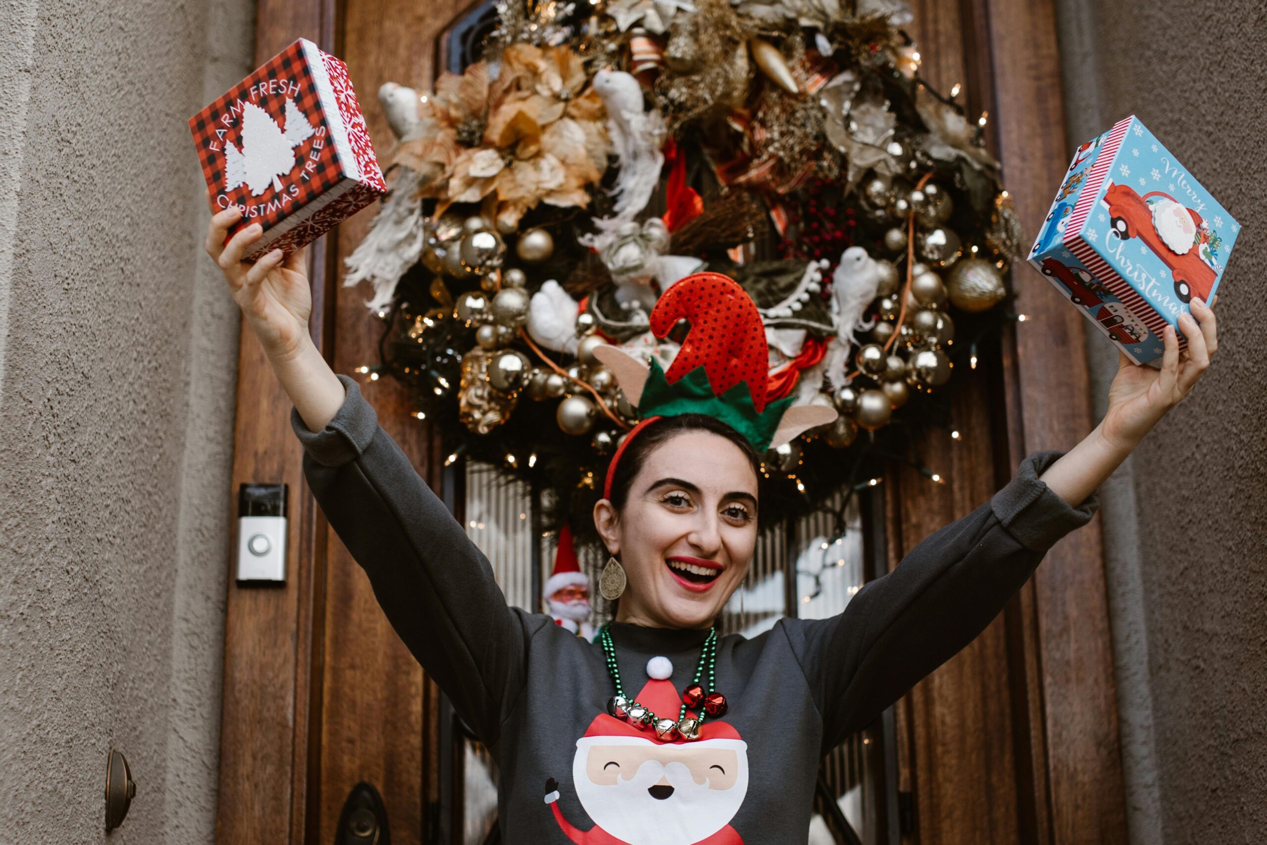 Joyful woman in an elf hat holding Christmas gifts by a decorated front door wreath.