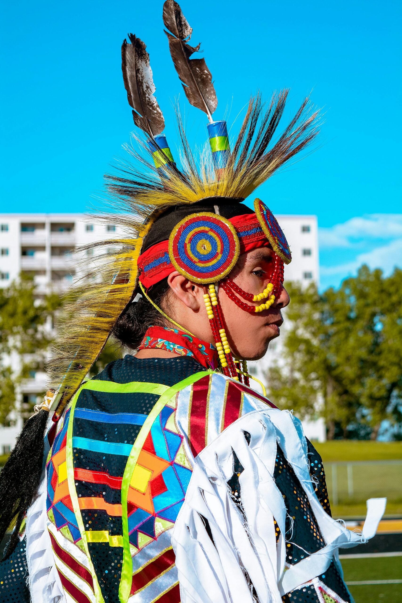 A Native American man in colorful traditional attire stands outdoors, showcasing cultural heritage.