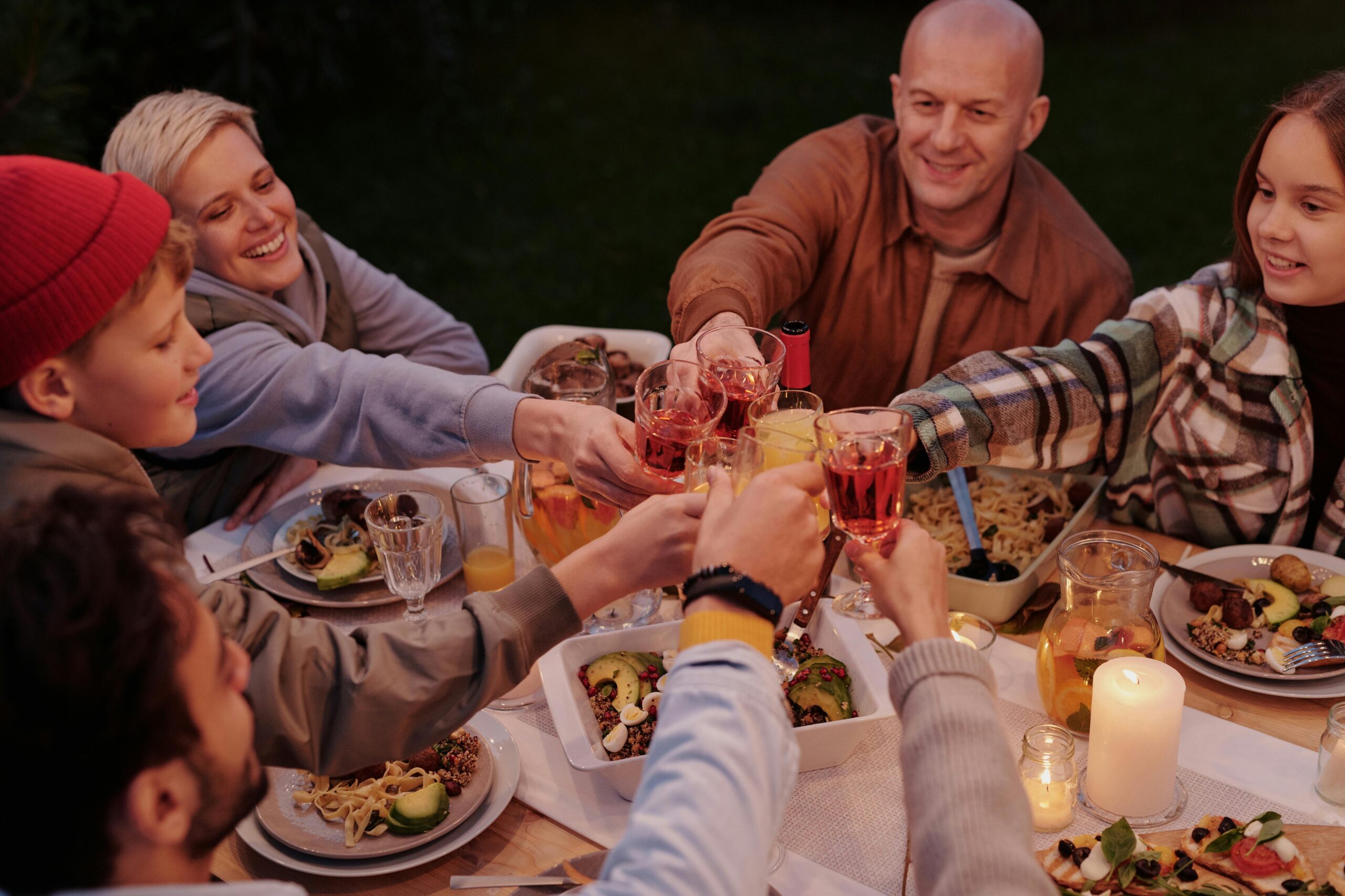 Smiling adults with children clinking with glasses while spending evening at dinner table in dusky garden